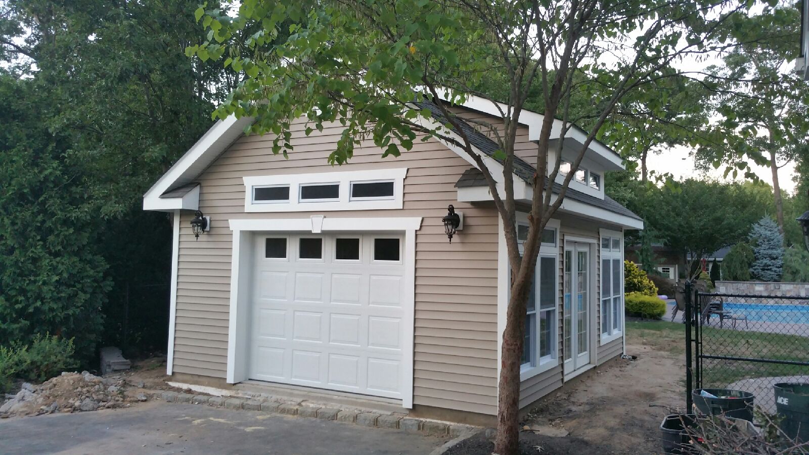 Custom detached garage with tan lap siding, white trim, carriage-style garage door, and double side doors amidst backyard greenery